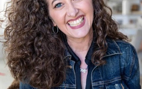Edie headshot wearing a denim jacket and smiling at camera with long dark wavy hair
