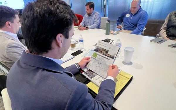 Man in a suit holds a marketing flyer while sitting at a table with other professionals in a conference room.
