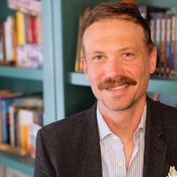 Joshua Fletcher's headshot showing him in a dark suit with a teal bookcase filled with books