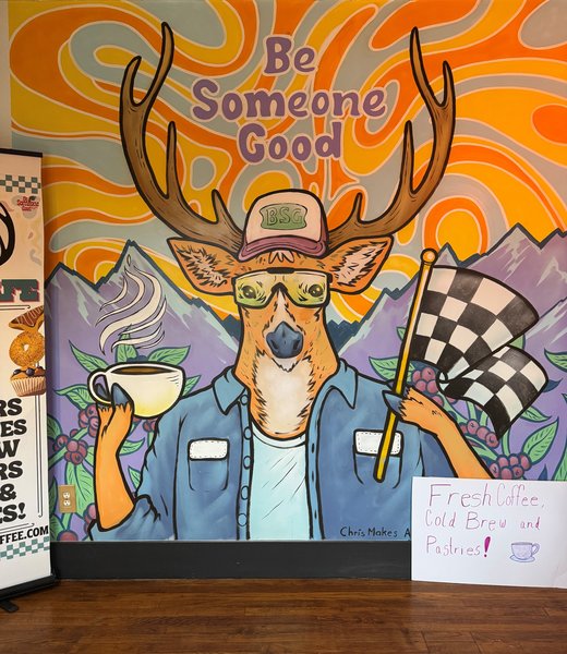 Mural with a deer wearing a hat and holding a cup of coffee and a checkered flag. Painted in purple it reads 'be someone good'